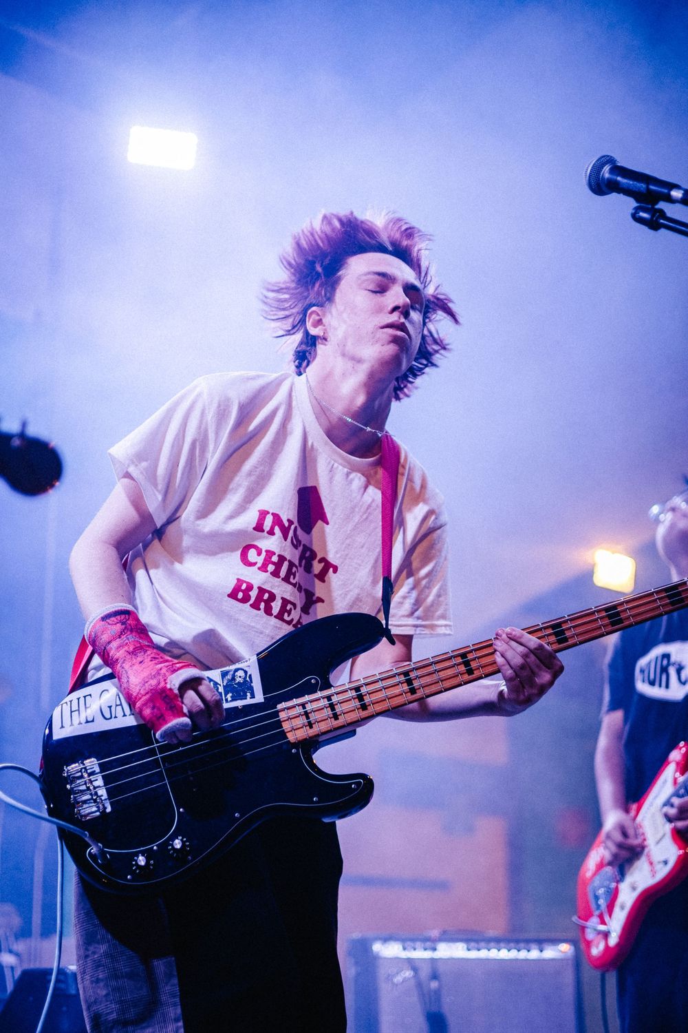 Energetic musician plays bass guitar on stage with dramatic blue and purple lighting during a live performance.