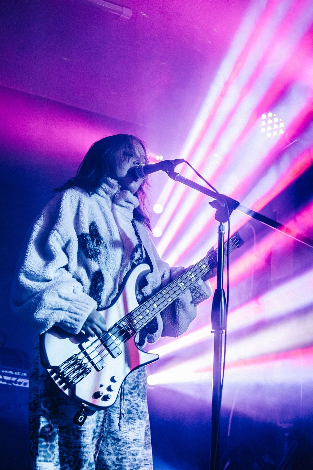 Bass guitarist performing on stage with dramatic pink and purple concert lighting and microphone setup.
