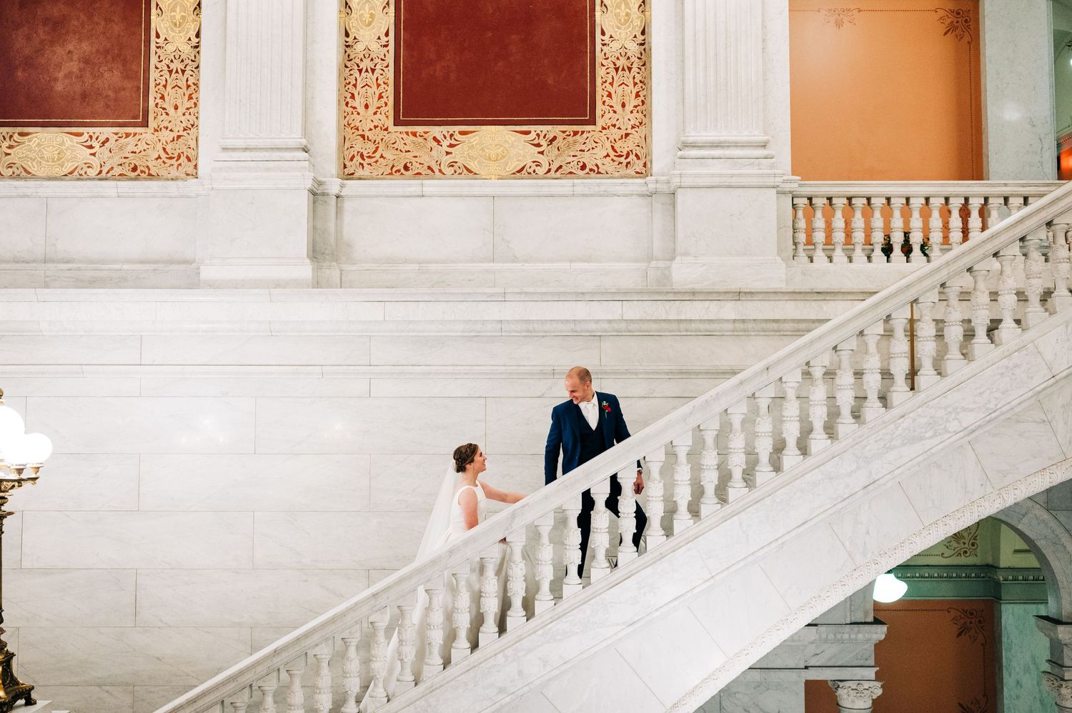 A couple descends an elegant white marble staircase in a historic building with ornate walls and classical architecture.
