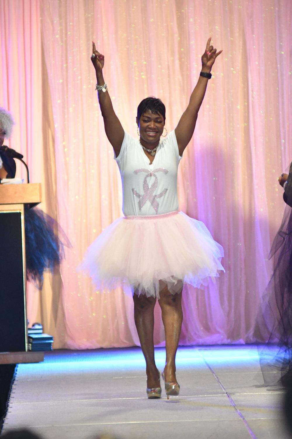 Joyful model in white tutu dress raises arms triumphantly on pink-lit runway.