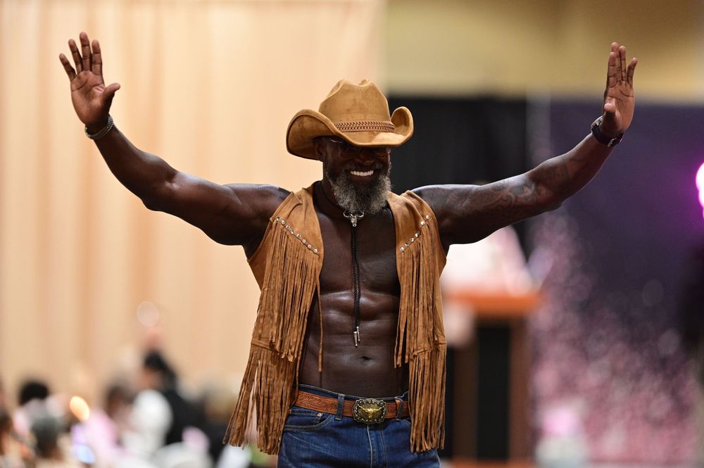Muscular performer in cowboy attire with fringed leather vest and hat raises arms on stage during show.