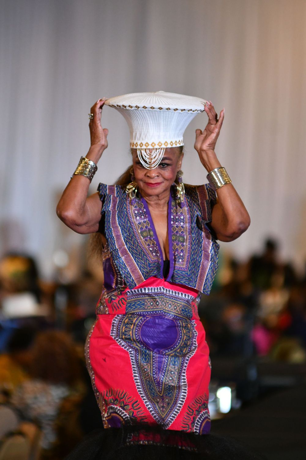 Model wearing colorful traditional African dashiki dress balancing decorative white bowl on head during fashion show.