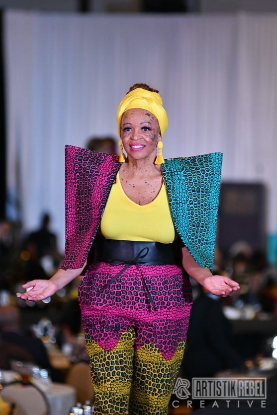 Model in colorful printed outfit with yellow headwrap poses on fashion runway.