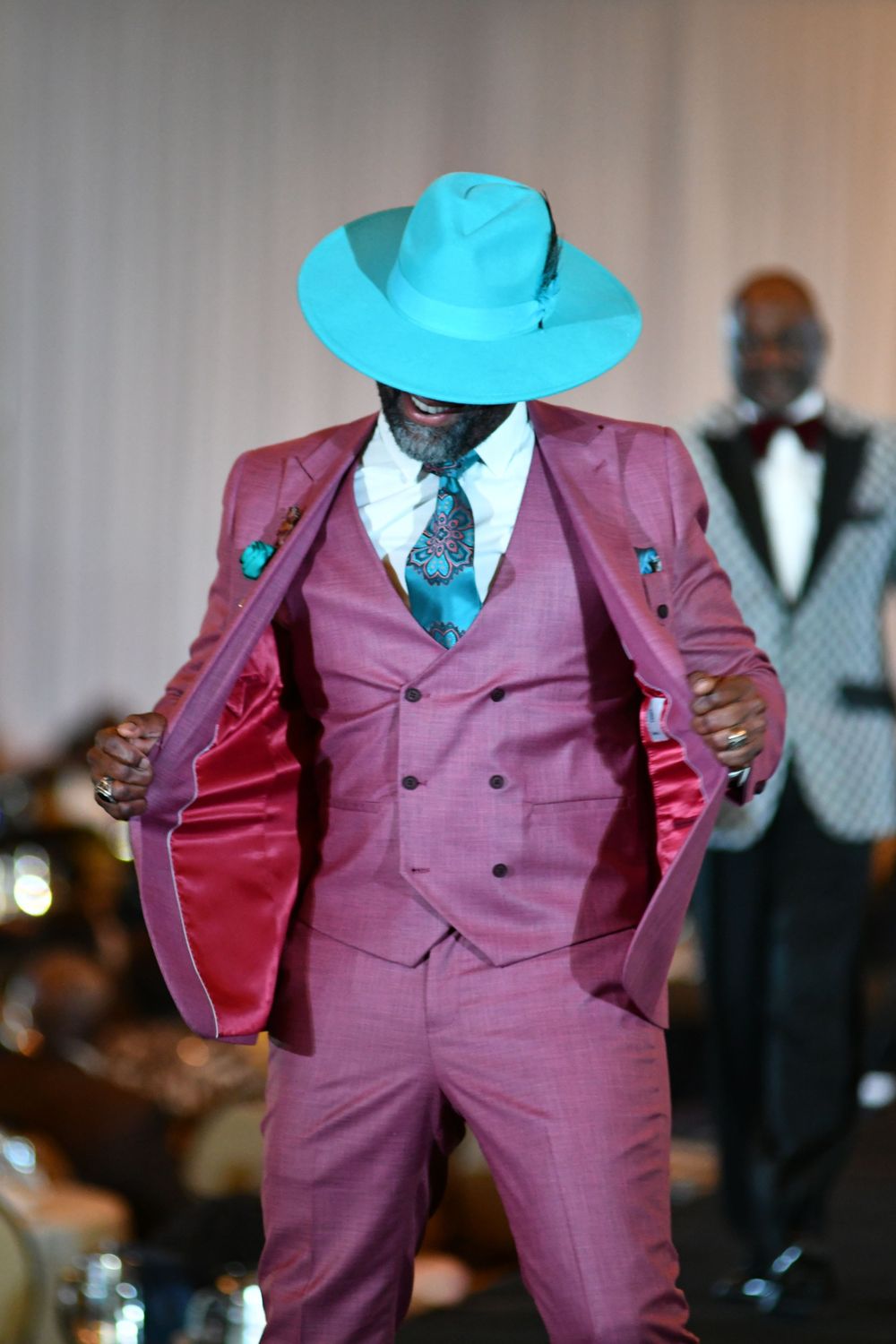 Model in mauve three-piece suit and turquoise hat striking pose on runway.
