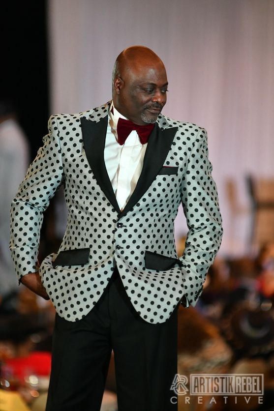 Model wearing polka dot tuxedo jacket with bow tie during fashion show.