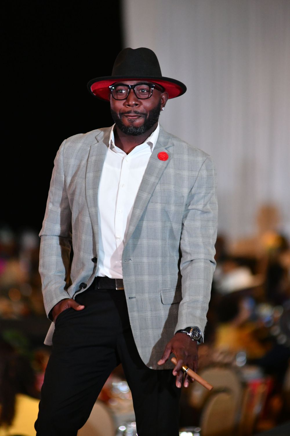 Stylish model wearing gray checkered blazer and red hat poses on fashion runway.
