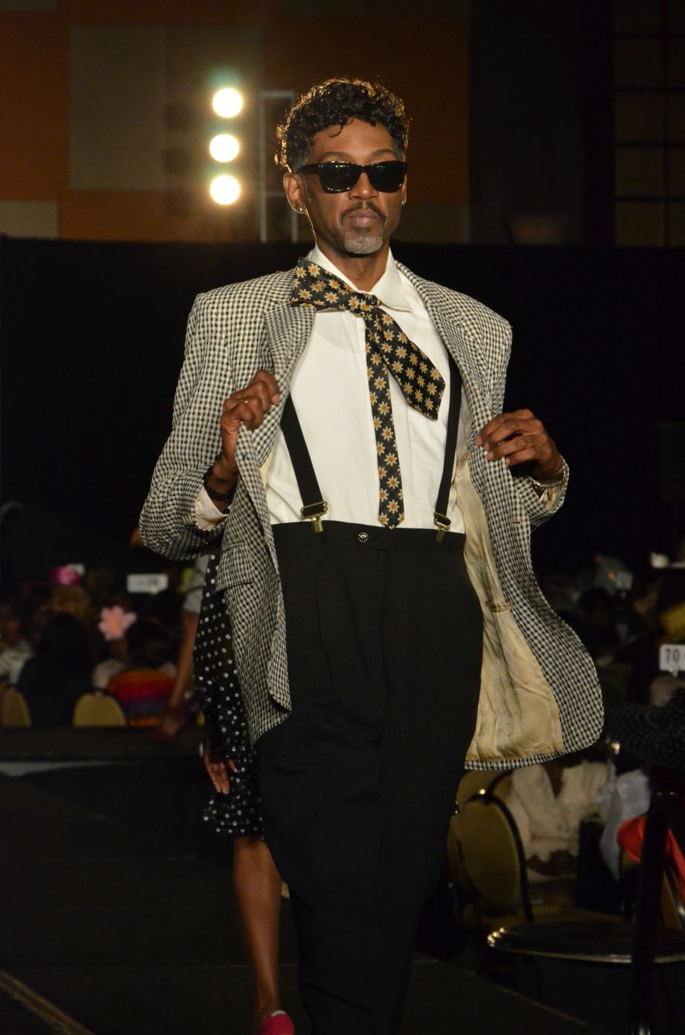 Stylish model in checkered blazer, suspenders and bowtie strikes pose on catwalk.