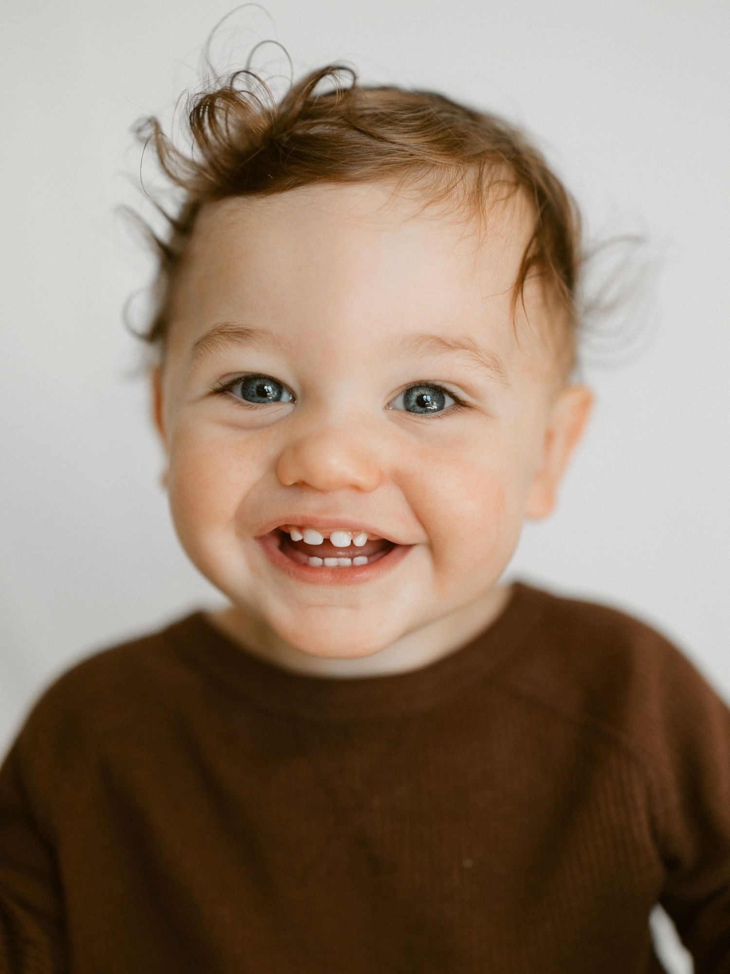 A smiling infant wearing a sage green onesie lies on a white textured blanket with brown flower petals nearby.