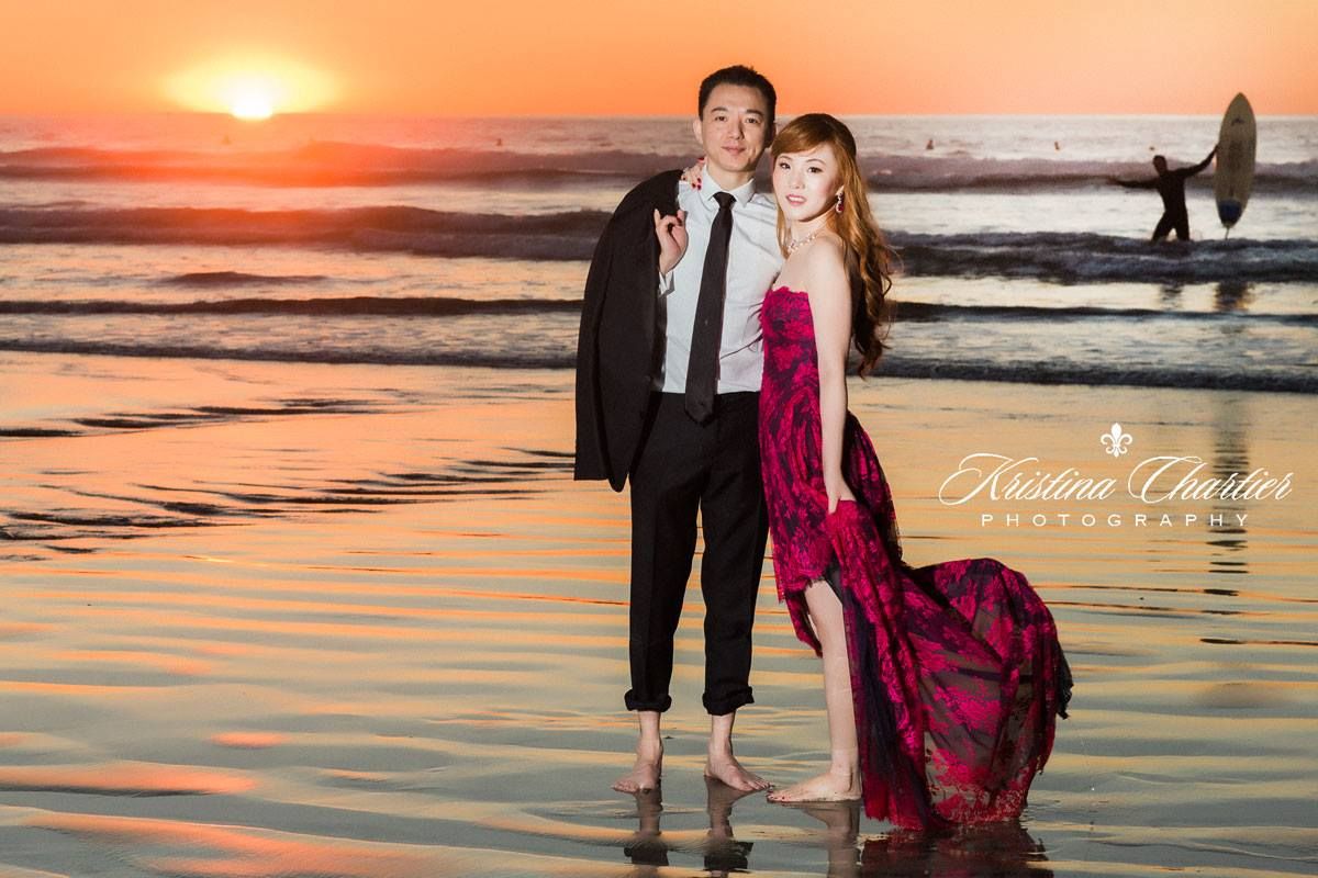 A couple poses on a beach at sunset with rippling water reflections and a flowing burgundy dress in the foreground.