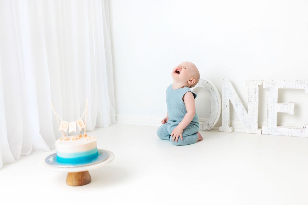 Baby in blue romper sits beside white ONE letters and birthday cake during first birthday photo session.