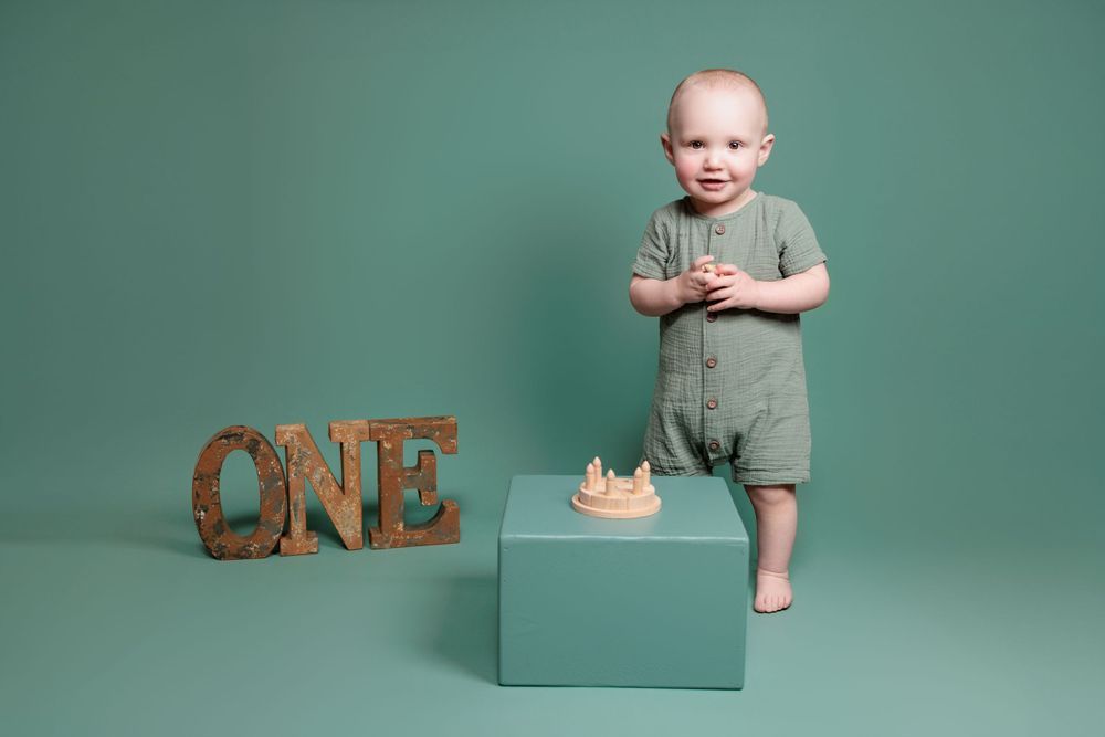Baby in sage green romper stands on blue platform next to wooden 'ONE' letters and gold crown in studio setting.