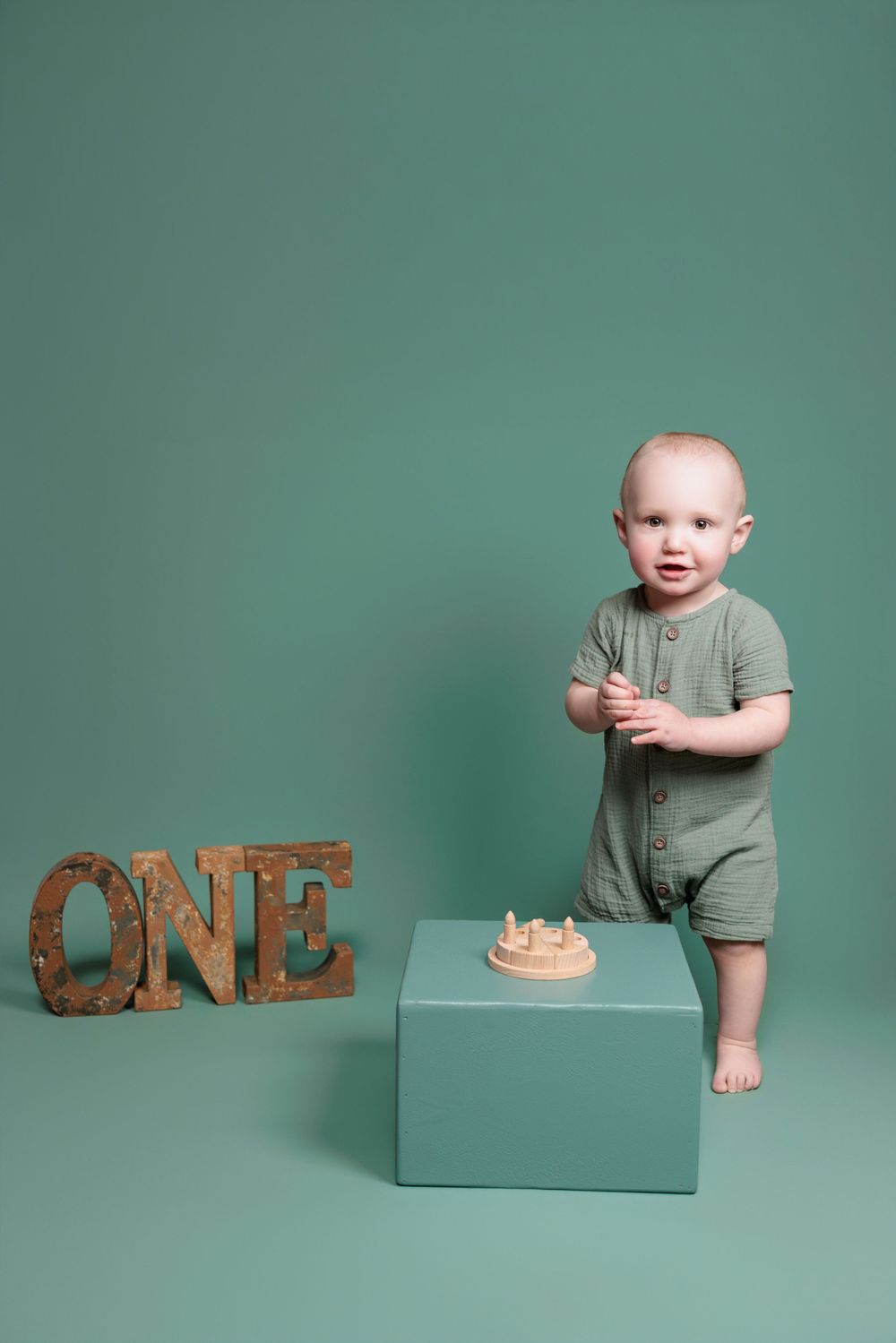 Baby in sage green romper stands on blue box next to decorative ONE sign and birthday cake against green backdrop.