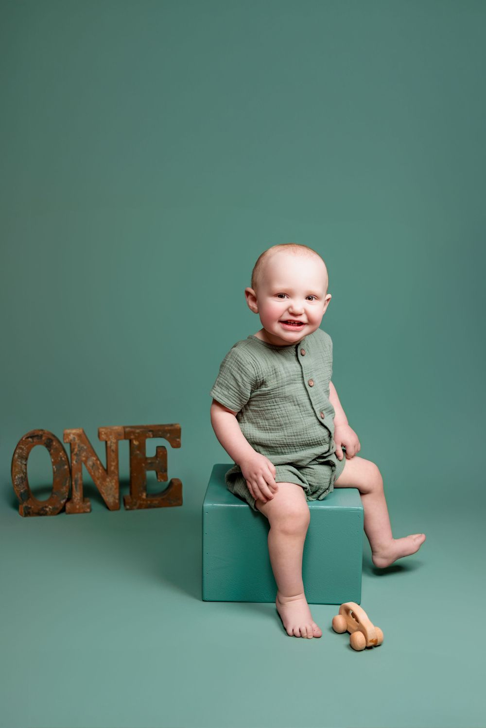 Baby in sage green outfit sits on turquoise box next to wooden 'ONE' sign and toy against teal backdrop.