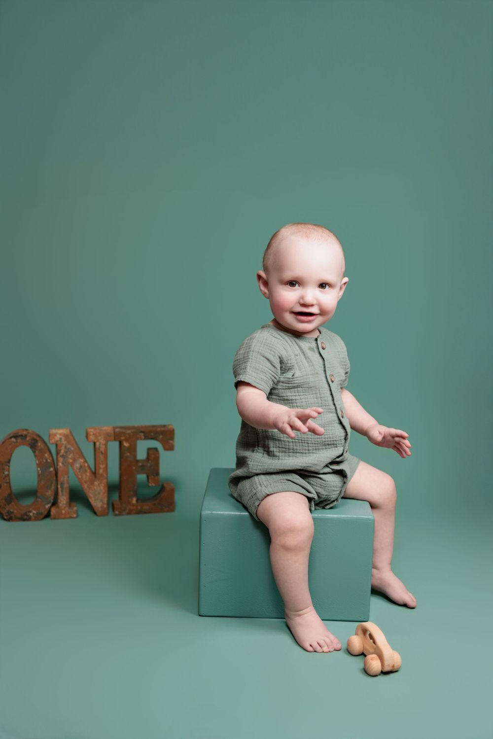 Baby in sage green romper sits on turquoise block next to wooden ONE letters against teal backdrop.