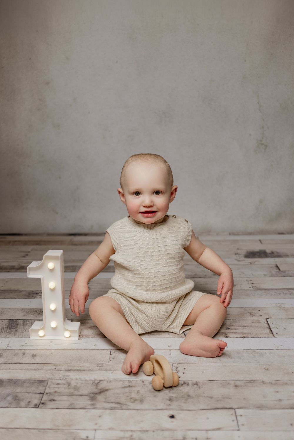 Baby in neutral-colored outfit sits on wooden floor next to illuminated number one and wooden toy.