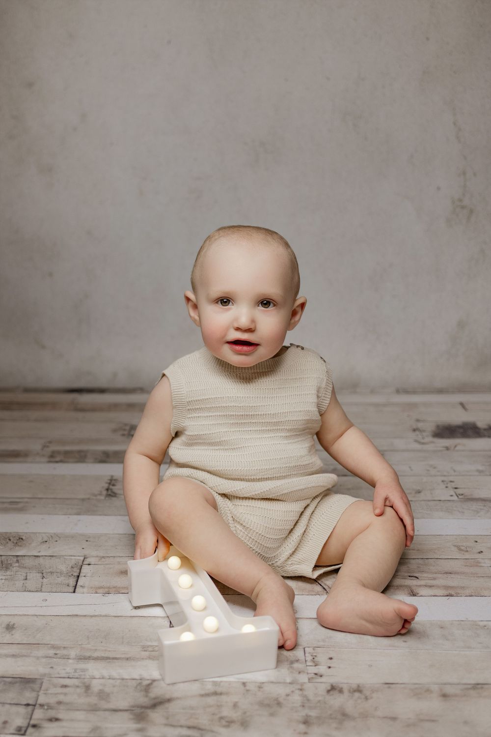 Baby in cream romper sits on wooden floor next to illuminated number one.