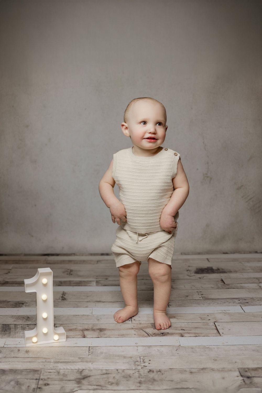 Small toddler in cream-colored outfit standing on wooden floor next to illuminated number one decoration.