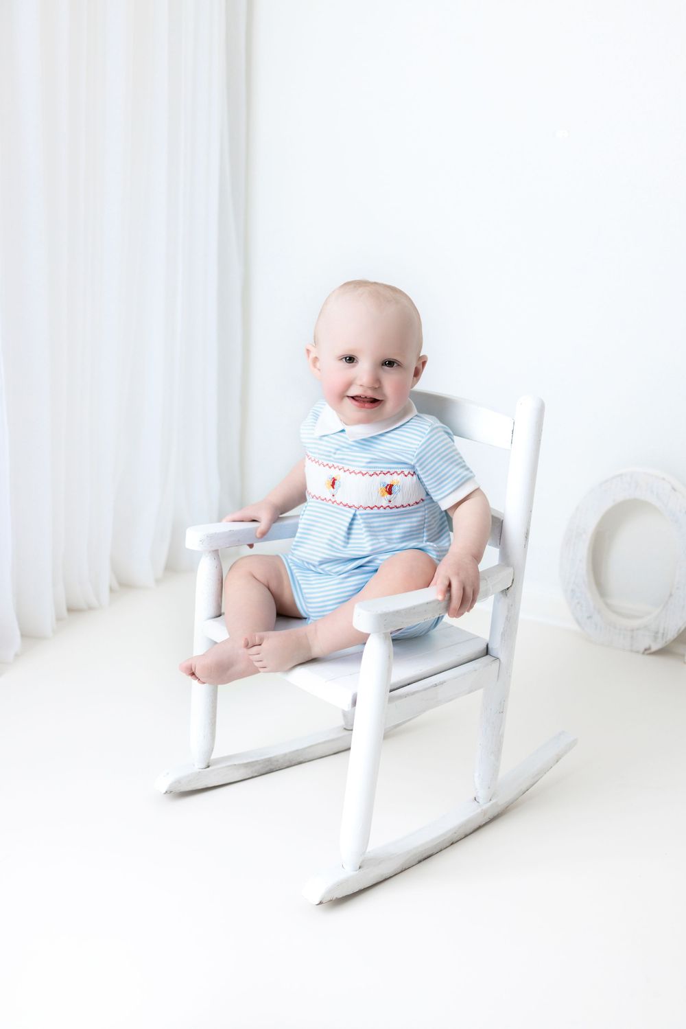 A smiling baby in light blue smocked outfit sits in a white wooden rocking chair against a bright white backdrop.