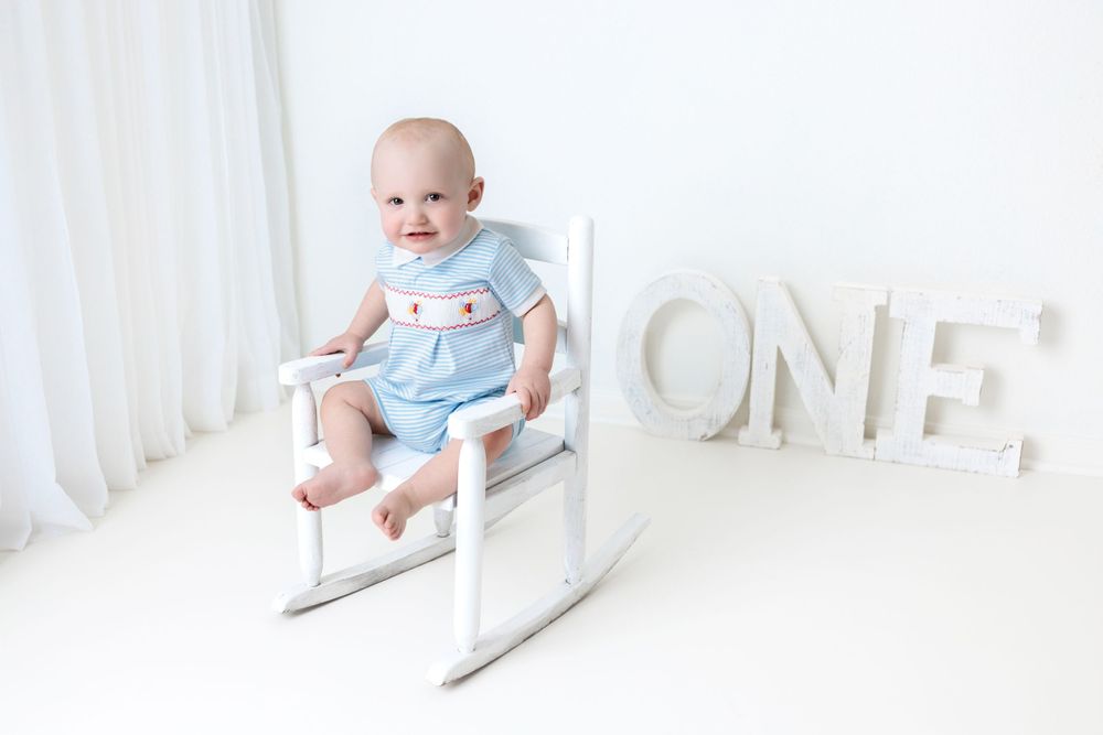 A baby in light blue clothes sits in a white rocking chair next to large white 'ONE' letters.