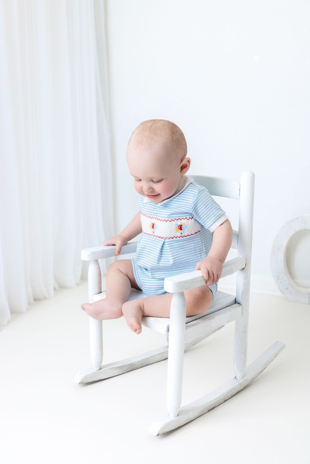Baby in light blue smocked outfit sits in white rocking chair against bright white background.
