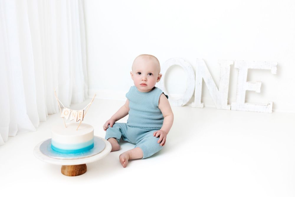 A baby in blue outfit sits next to white 'ONE' letters and a smash cake for first birthday photoshoot against white backdrop.