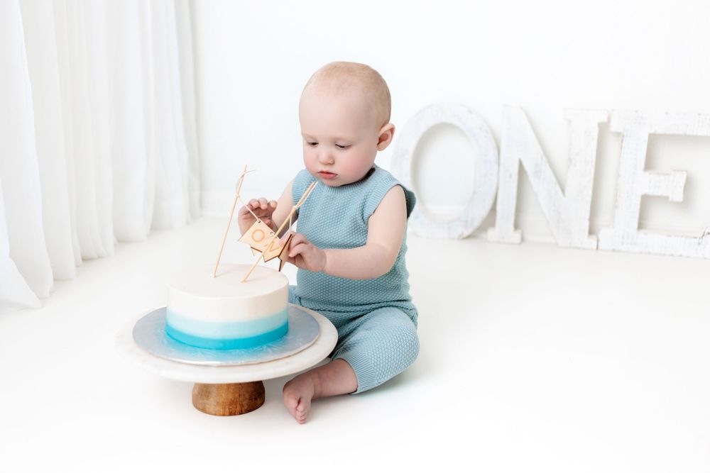 Baby in teal romper sits on floor with birthday cake and decorative 'ONE' letters against white backdrop.