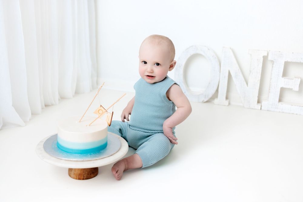 Baby in blue outfit sits next to birthday cake and white ONE letters against light background.
