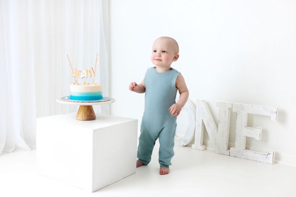 Baby in teal romper stands next to birthday cake and white ONE letters in minimalist setting.