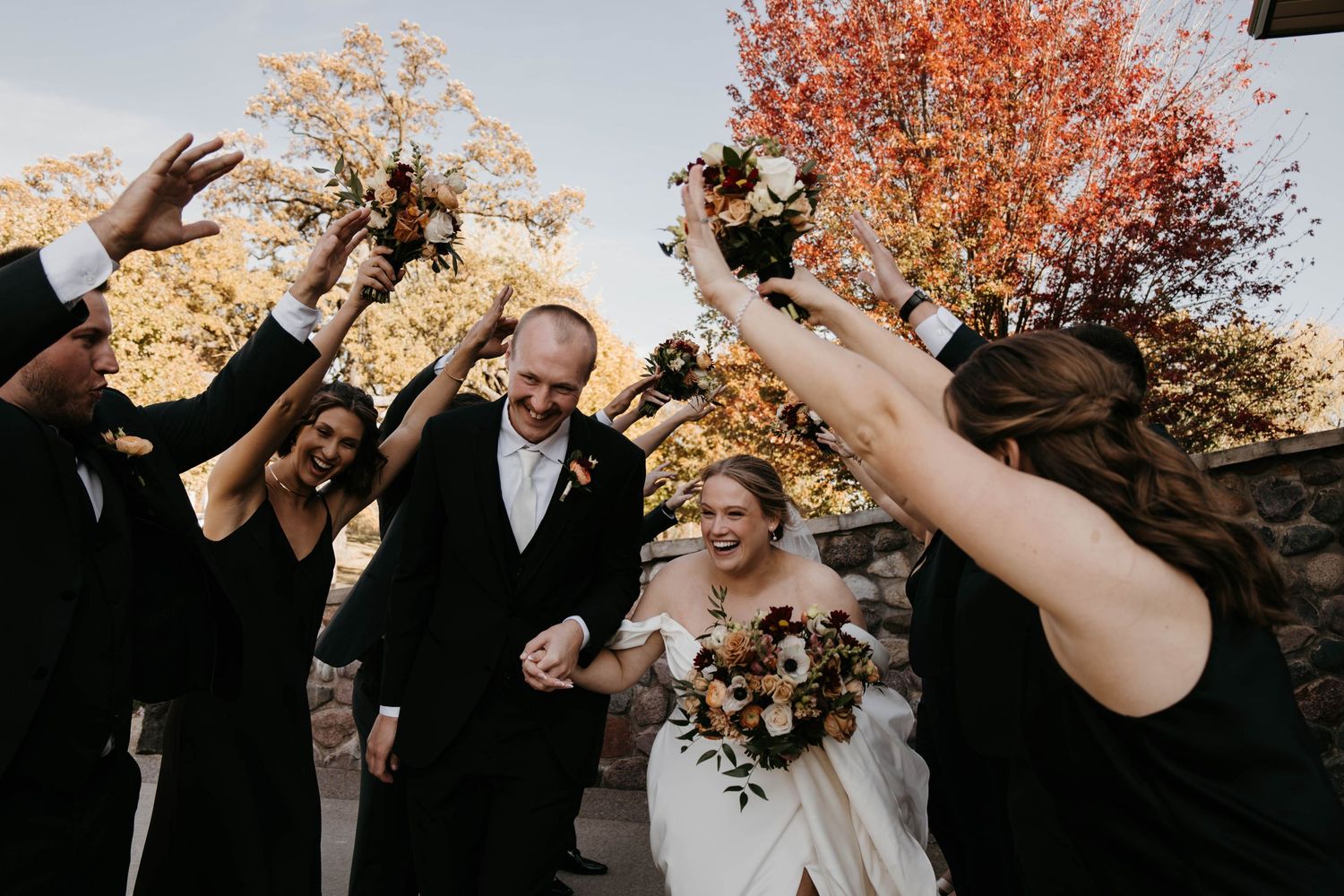 Bride and groom walk through cheering wedding party amid autumn foliage at outdoor fall wedding ceremony.