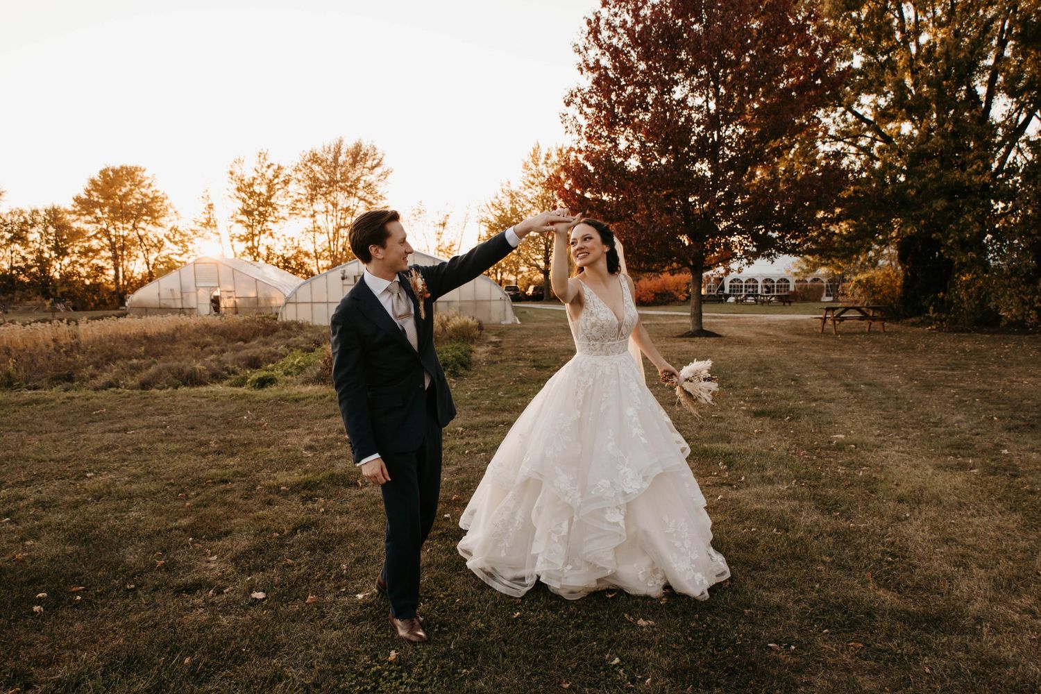 Bride and groom dance in golden autumn light on a grassy field, with a white tent venue in the background.