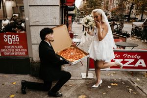 Groom kneels presenting open pizza box to laughing bride on city street after wedding.