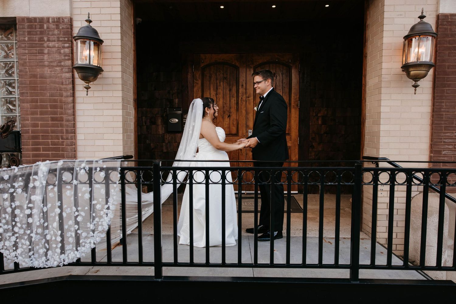 Bride and groom hold hands on ornate iron balcony outside brick church with cathedral doors.