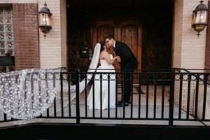 Bride and groom share romantic kiss on decorative balcony with sparkling veil cascading beside them.