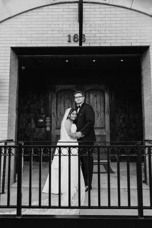 Black and white portrait of bride and groom embracing outside brick building entrance numbered 166.