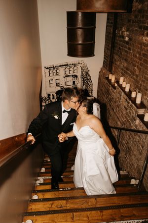 Bride and groom kissing on rustic wooden staircase with exposed brick and candlelit steps.