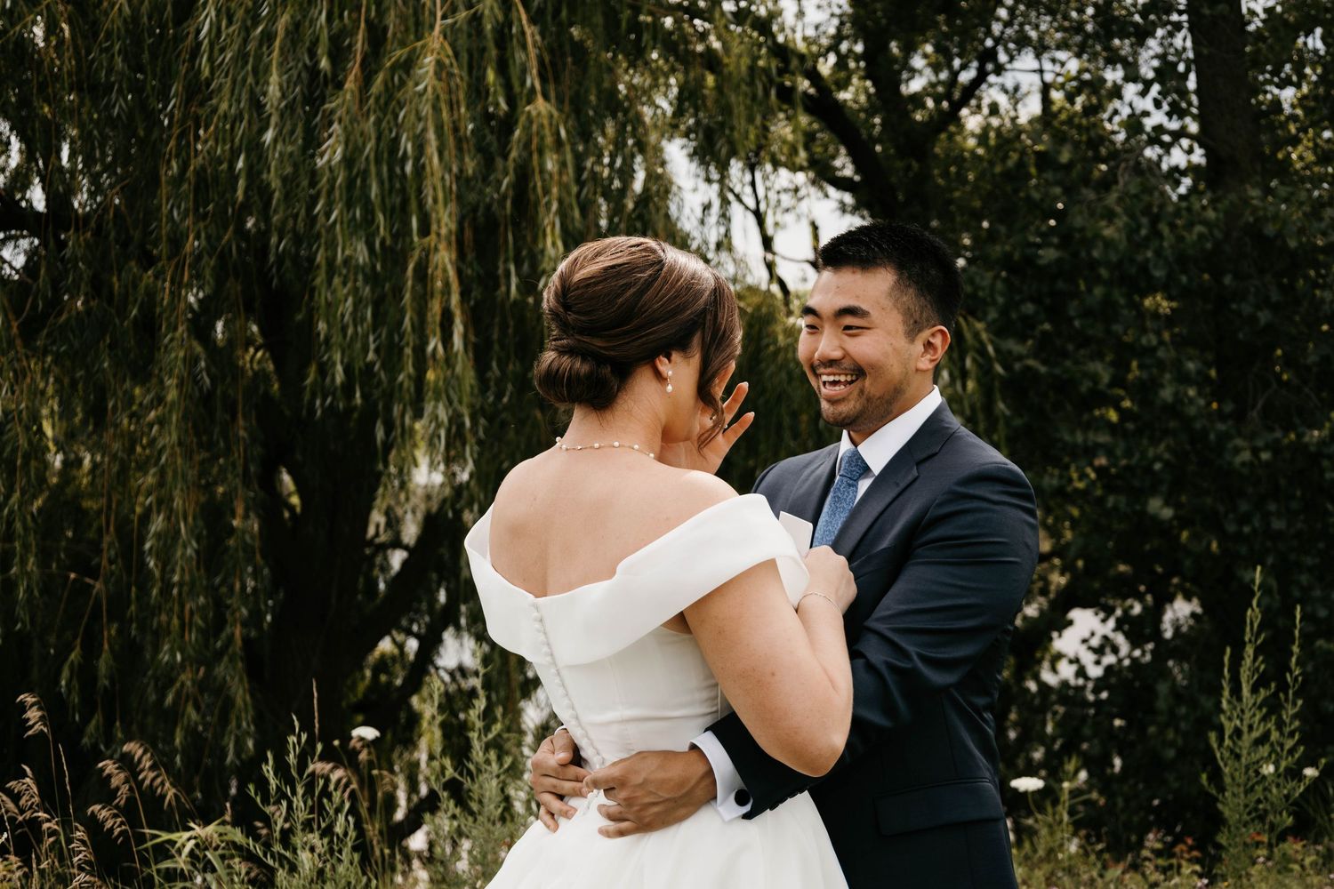 Smiling groom embraces bride in off-shoulder gown during first look near weeping willow tree.
