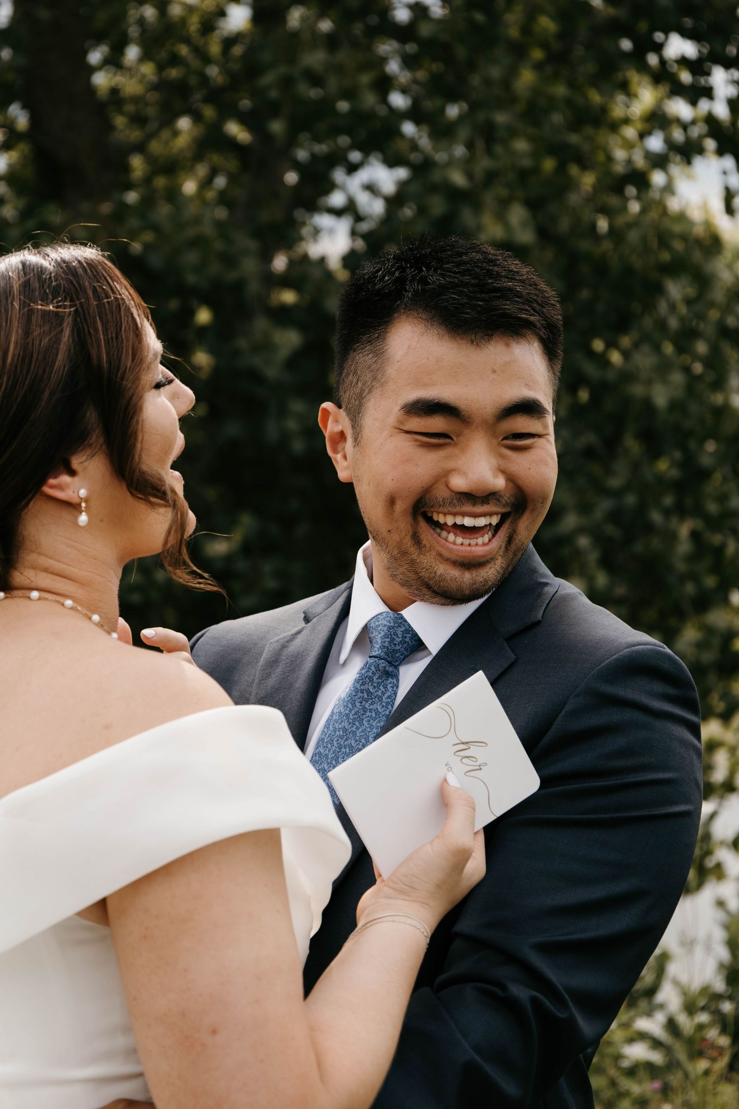 Joyful groom laughs while bride holds vows card during emotional first look moment outdoors.
