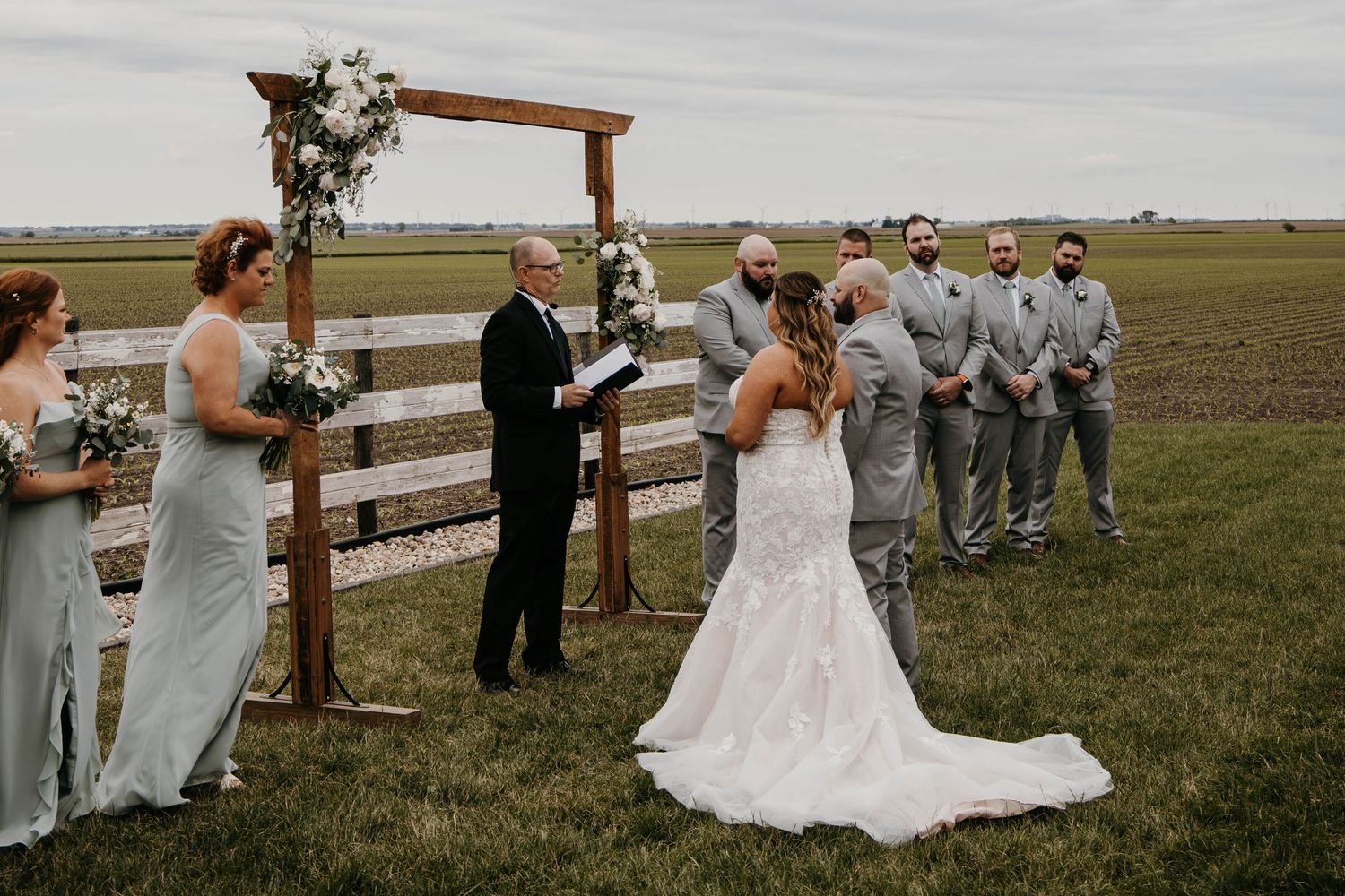 Bride and groom exchange vows at rustic wooden arch ceremony on open farm field.