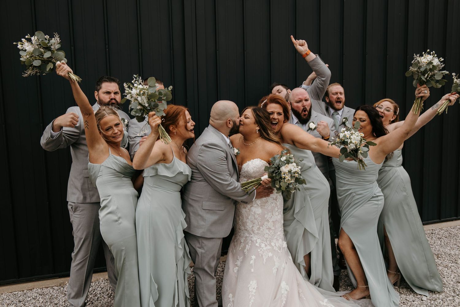 Excited wedding party surrounds kissing bride and groom in sage green and gray attire.