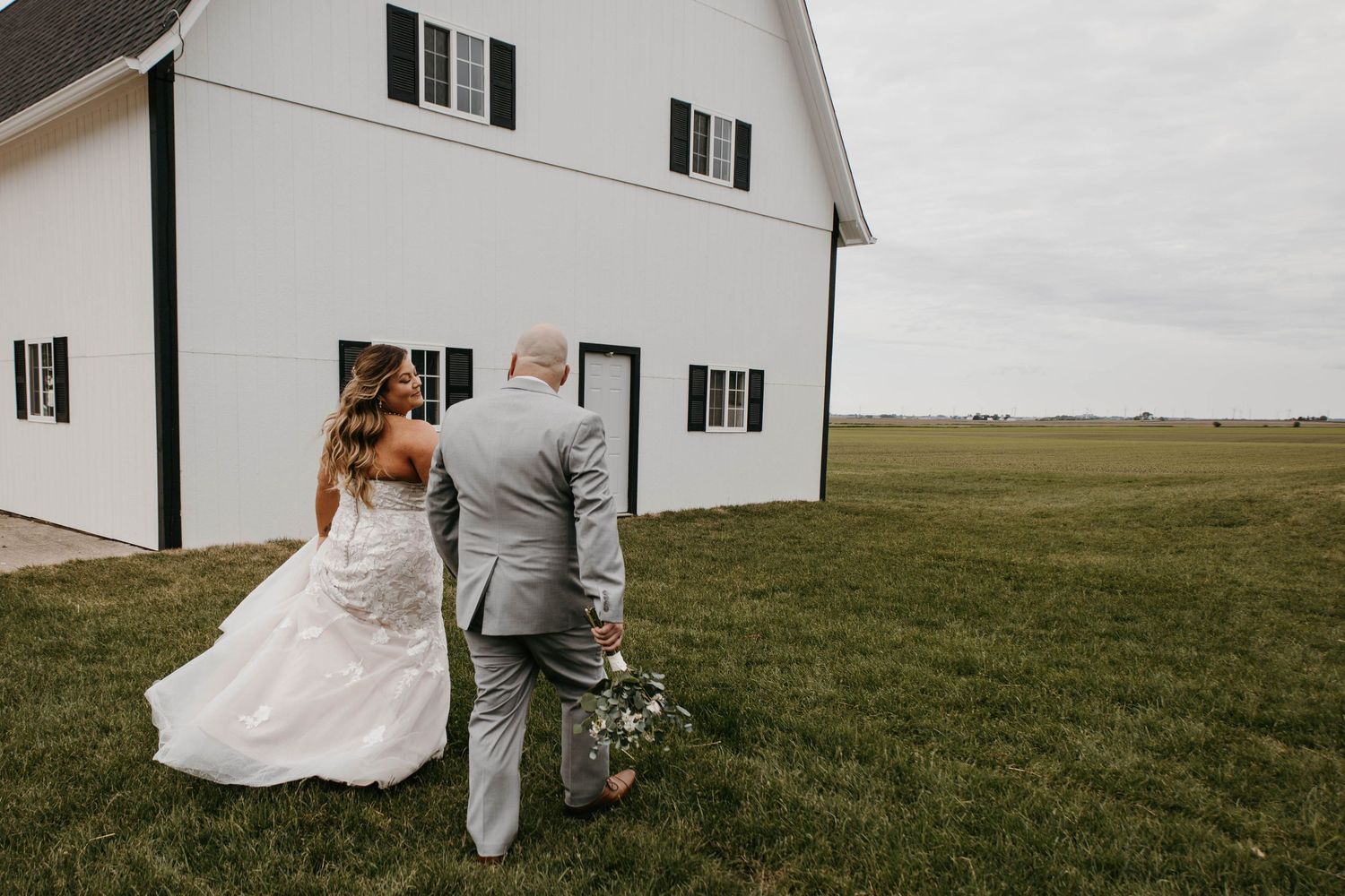 Bride and groom walk hand-in-hand toward white barn venue across lush green countryside.