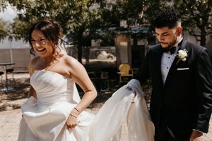Bride laughs as groom helps lift her white wedding gown outdoors on sunny day.
