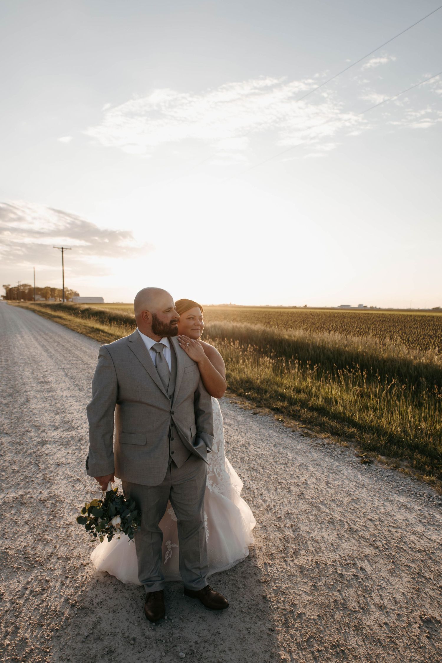 Bride and groom share golden hour kiss on rural gravel road surrounded by farmland.