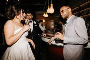Bride and groom clink champagne glasses with guest near bar at wedding reception.