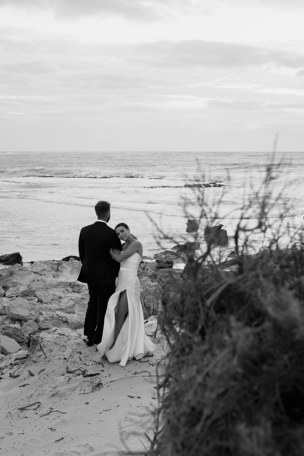 A romantic black and white photo of a couple embracing on rocky coastal cliffs overlooking the ocean.