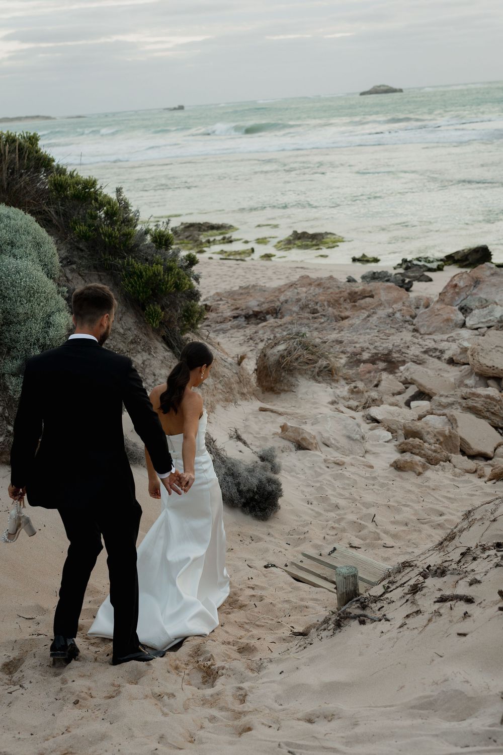 Couple in formal attire walking down rocky coastal path with ocean waves in background.