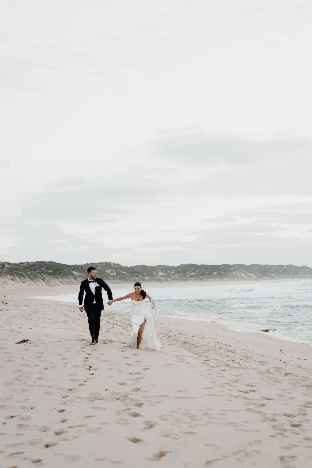 Newlyweds walking hand in hand along a windswept beach with sand dunes in the distance.