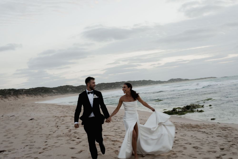 Joyful couple in wedding attire running together along a scenic beach at dusk.