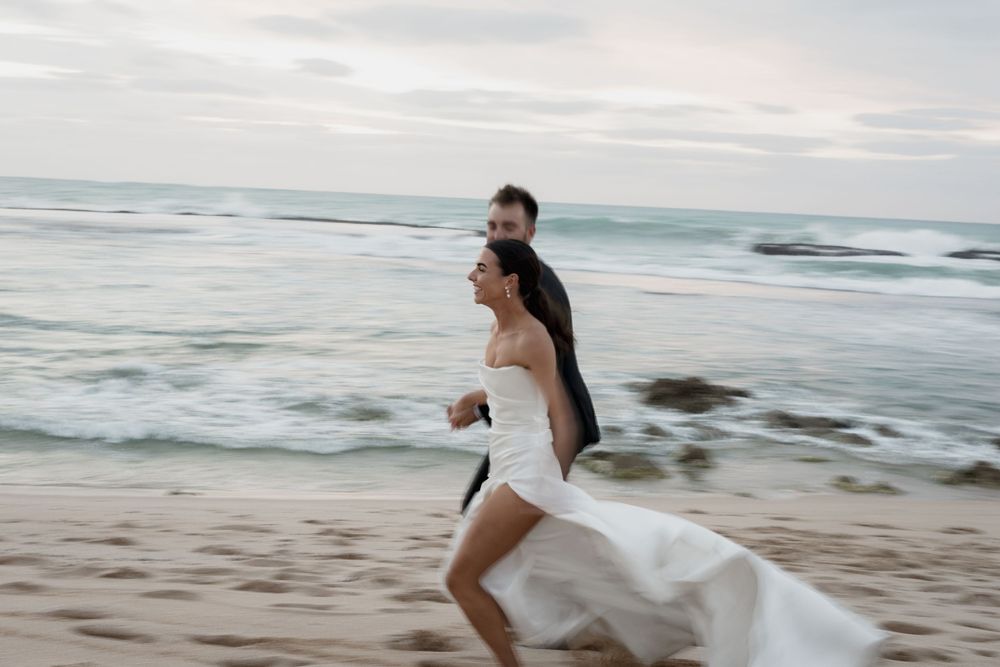 Happy couple in wedding attire running playfully on the beach as waves crash behind them.