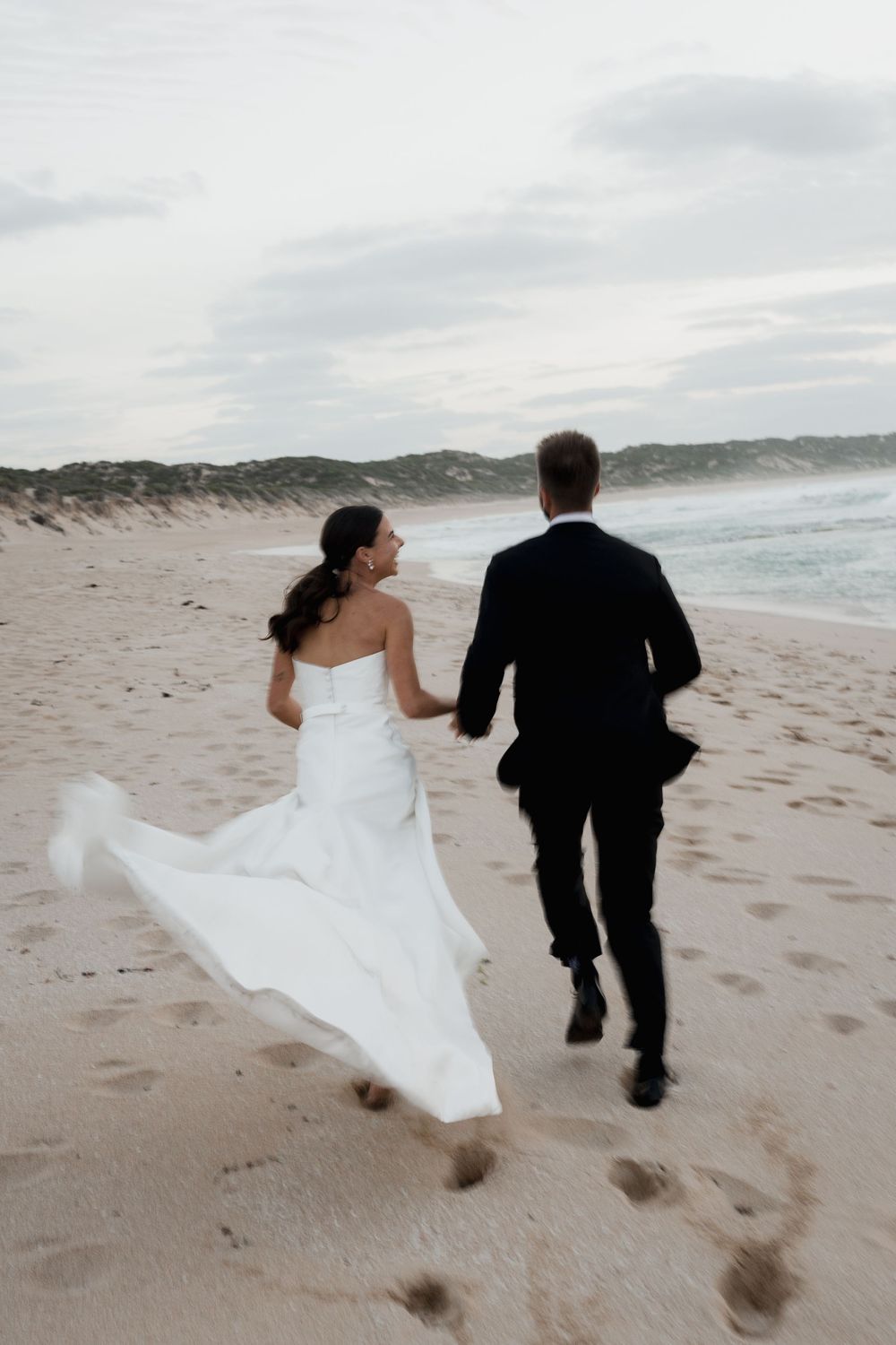 Couple in wedding attire walking along beach with flowing white dress catching the wind.