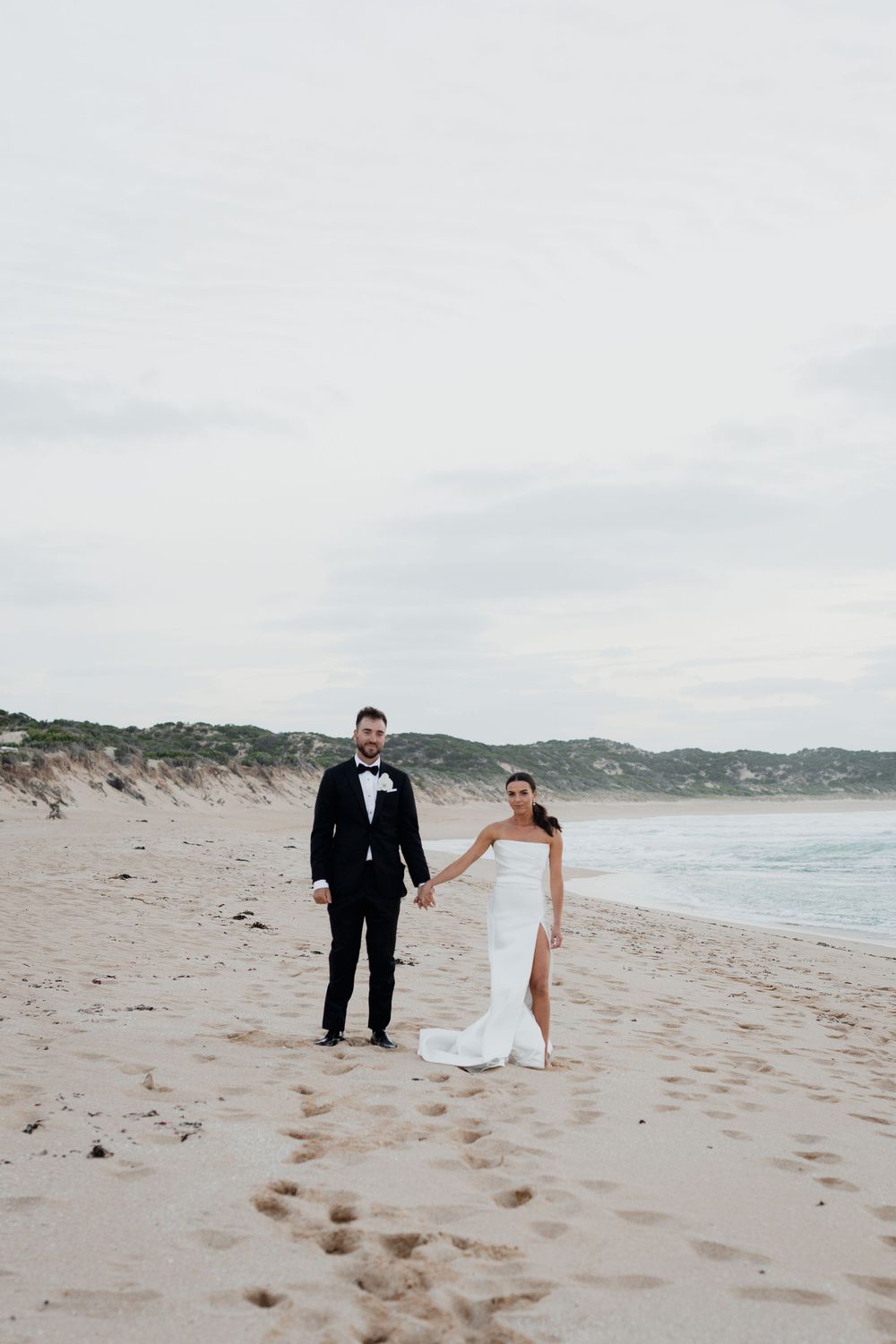 Elegant couple in wedding attire standing together on empty beach with coastal dunes in background.